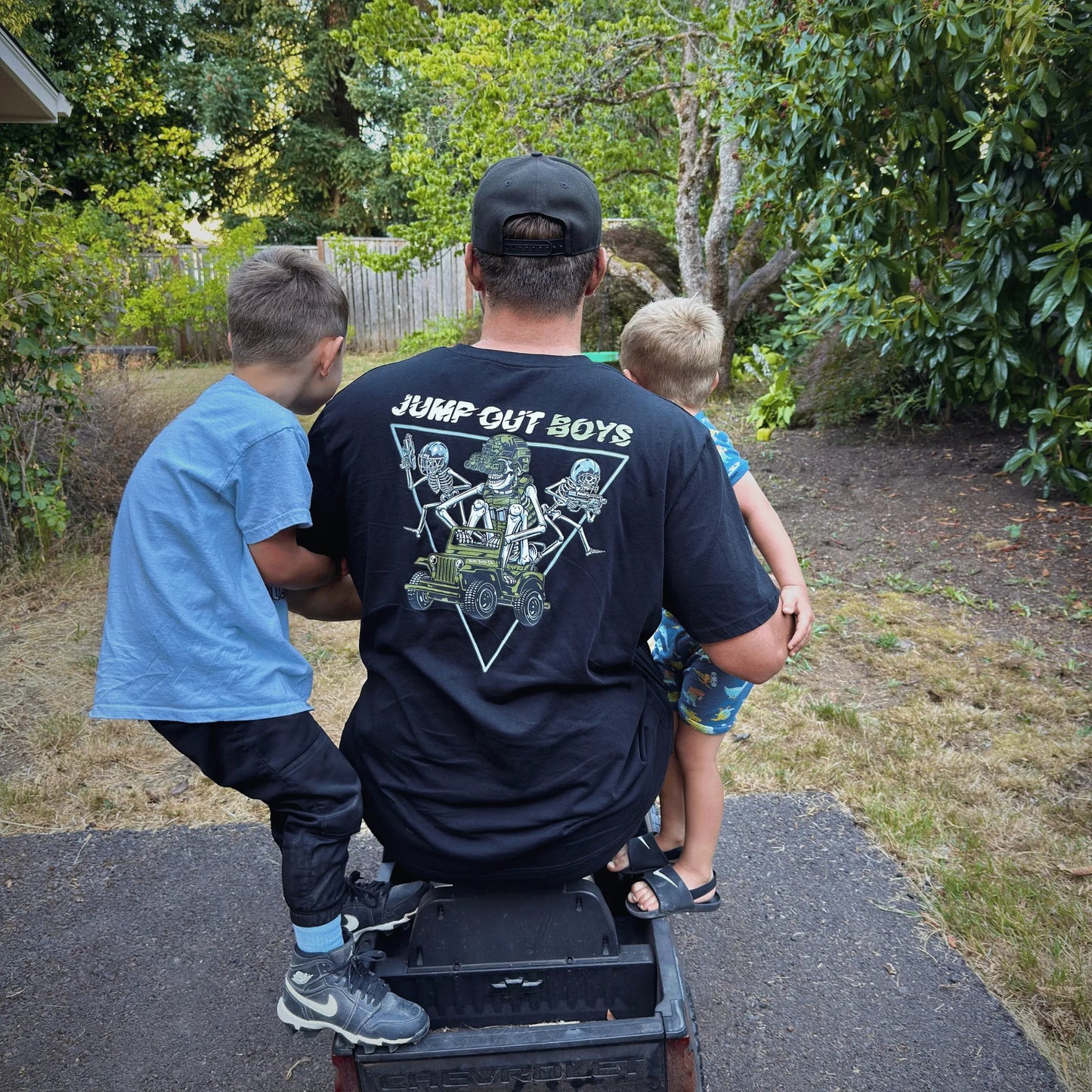 Dad wearing jump out boys shirt with 2 kids while on a power wheel